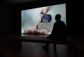 man sitting on a bench in a dimly lit gallery space watching a large scale projection of a nourishment can of drink being opened in water