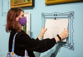 A visitor to Life with Art fixes their artwork to the blue wall - inside the drawing of an ornate frame
