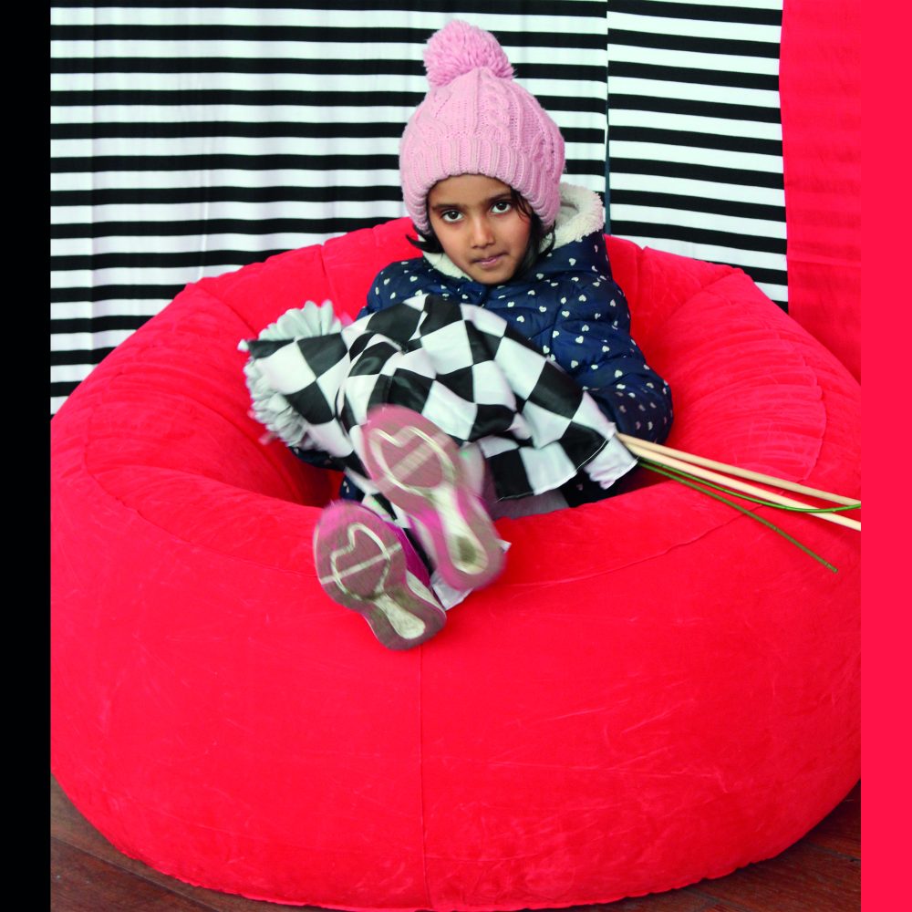 A young child sitting on a red bean bag holding a black and white checkered flag in front of a black and white stripe background.