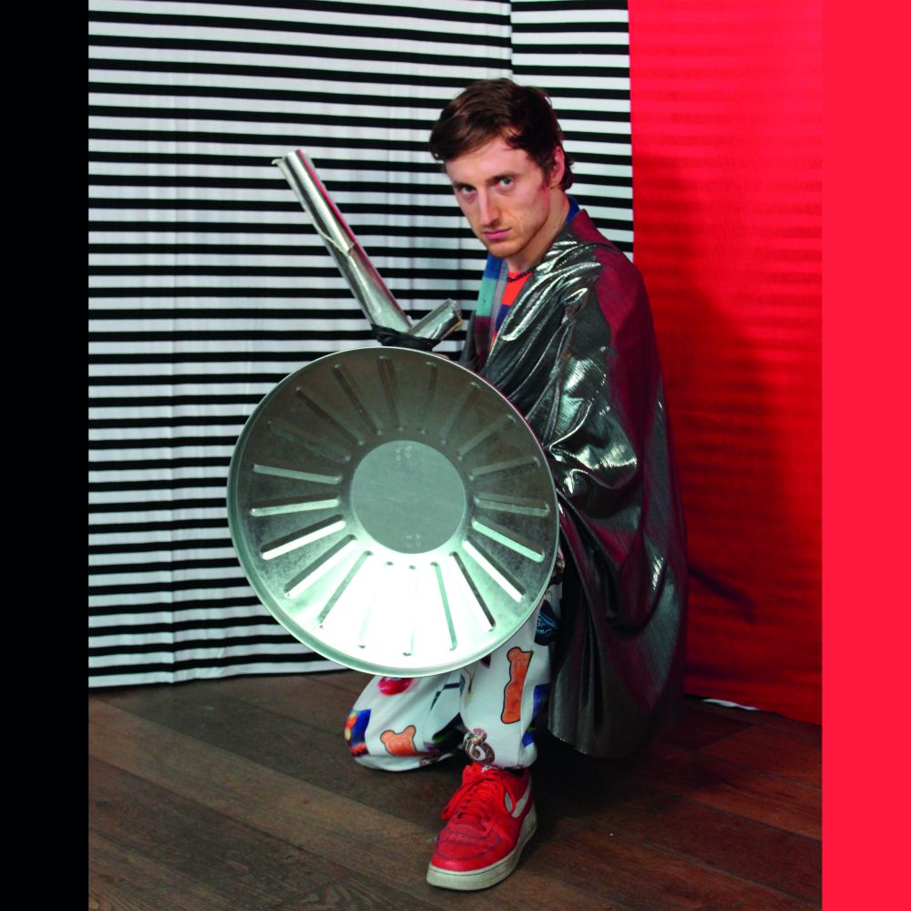 Man in a fancy dress costume holding a metal shield as he kneels on the floor in front of a red and black & white stripe background.