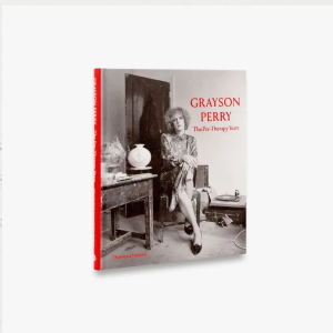 Cover of Grayson Perry: The Pre-Therapy Years has Grayson Perry dressed in a knee-length flower pattern dress and heals, sitting on a wooden chair next to a table covered with different objects. The image is in black and white.