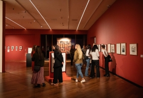 A wide shot of the main gallery at Firstsite, featuring red walls with paintings and drawings displayed next to each other. In the middle lots of visitors are looking at the artworks.