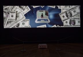 A wide screen projection of a moving image artwork showing a figure on a laptop screen, surrounded by dollar bills.