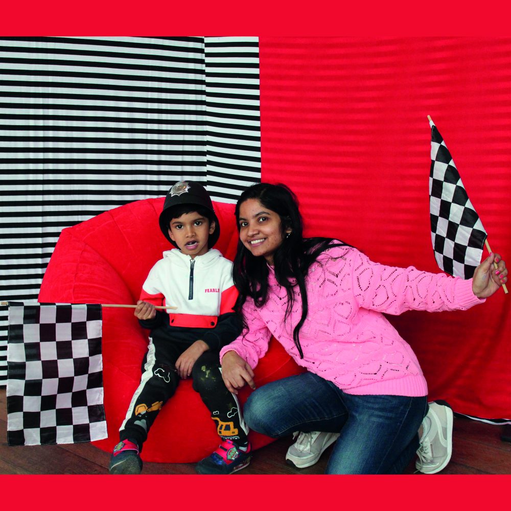 A woman kneeling in front of her young child whilst they sit on a red bean bag, both holding checkered flags in front of a red and black & white stripe background.