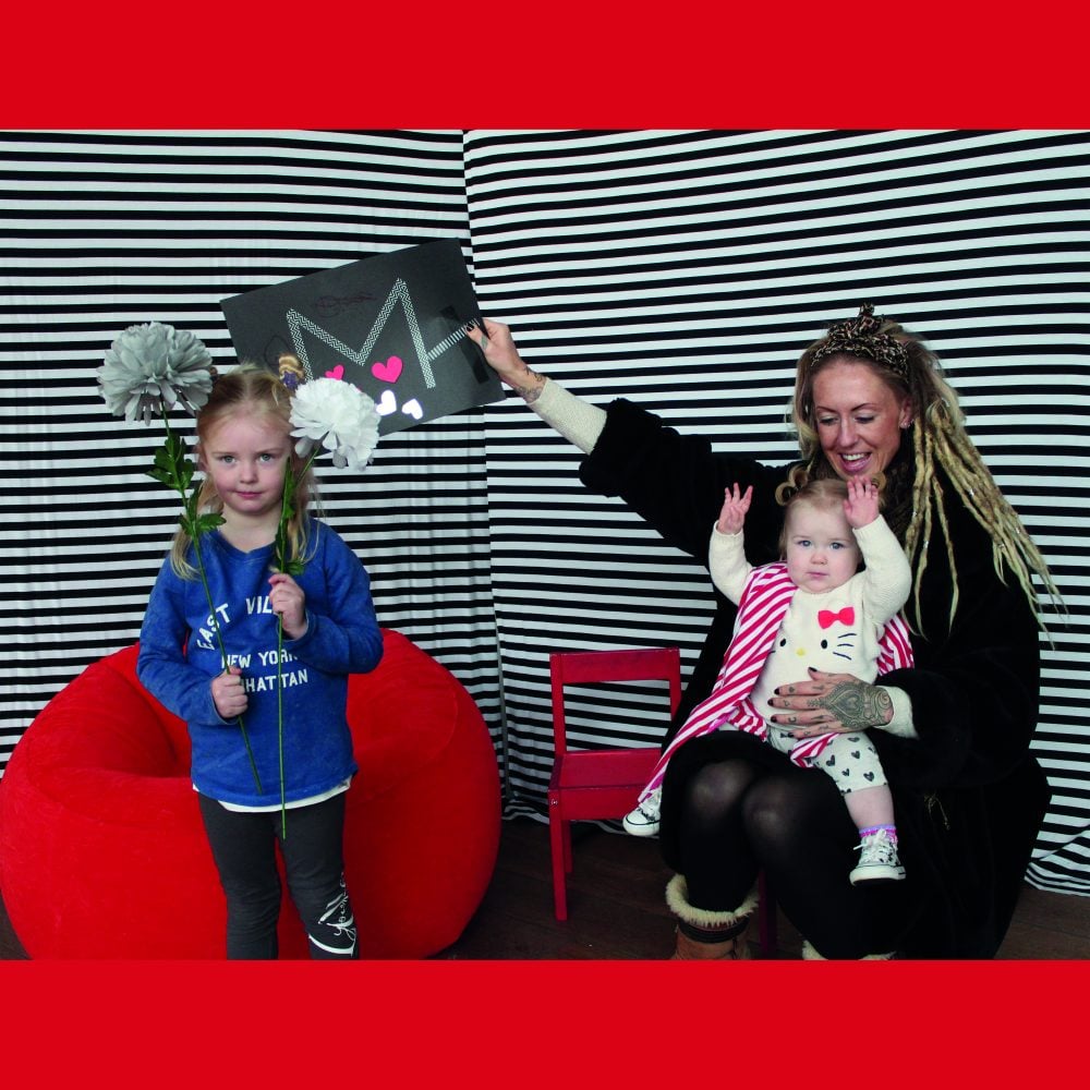 A woman and her two little children holding up a drawing in front of a red beanbag and chair in front of a black and white stripe background.