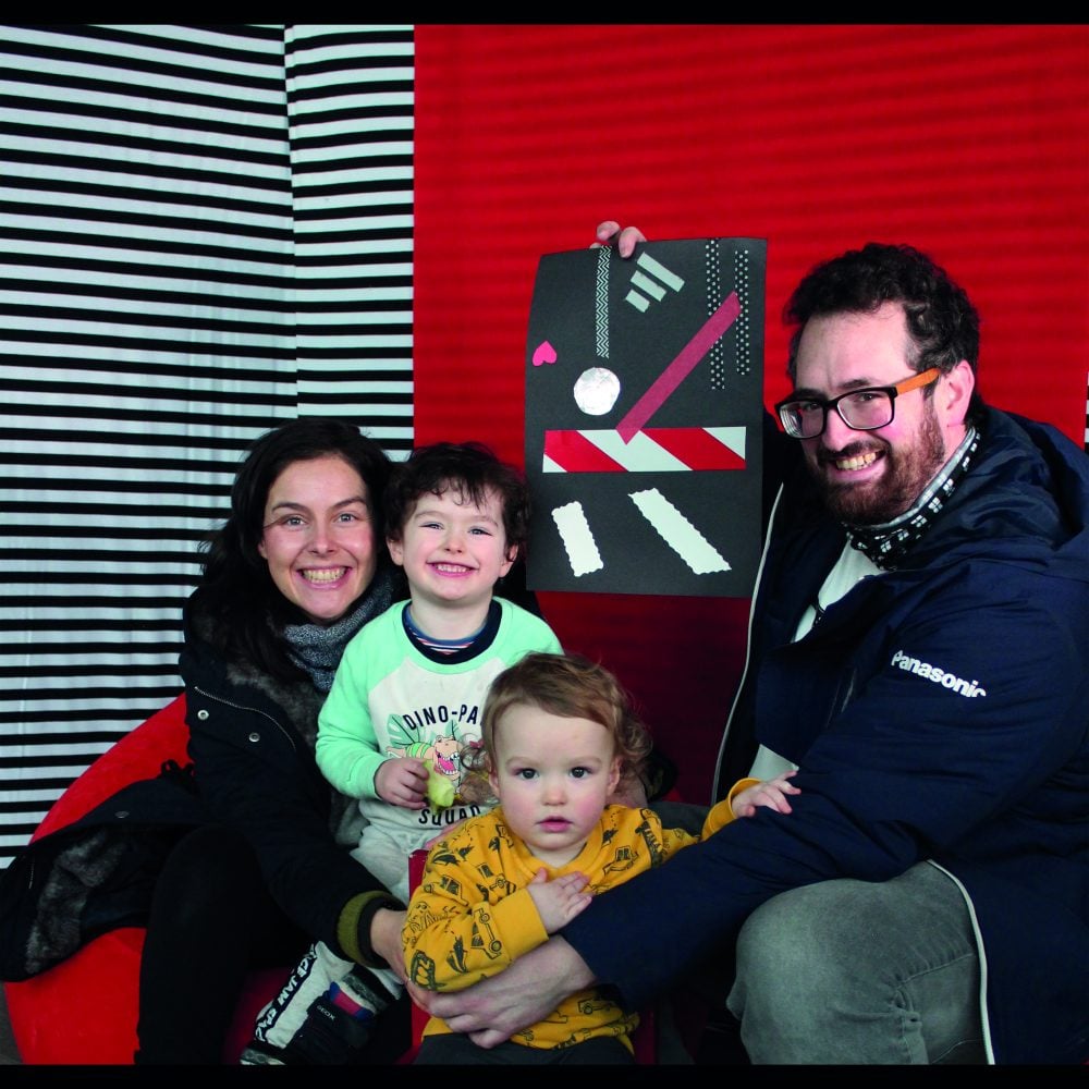 A man, a woman and their two little children sitting on a red bean bag holding up a drawing in front of a red and black & white stripe background.