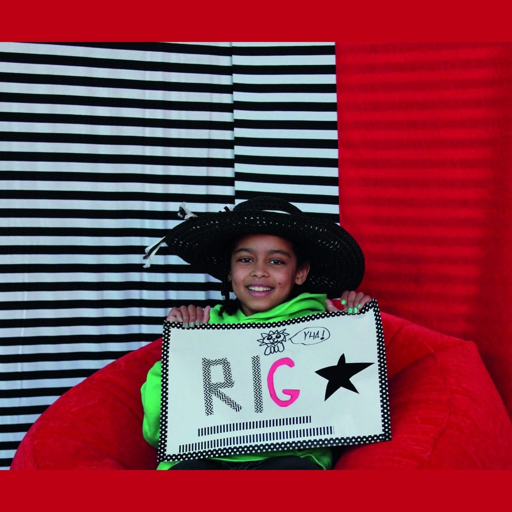 A child in a cowboy hat holding up a drawing on a red bean bag in front of a black & white stripe background.