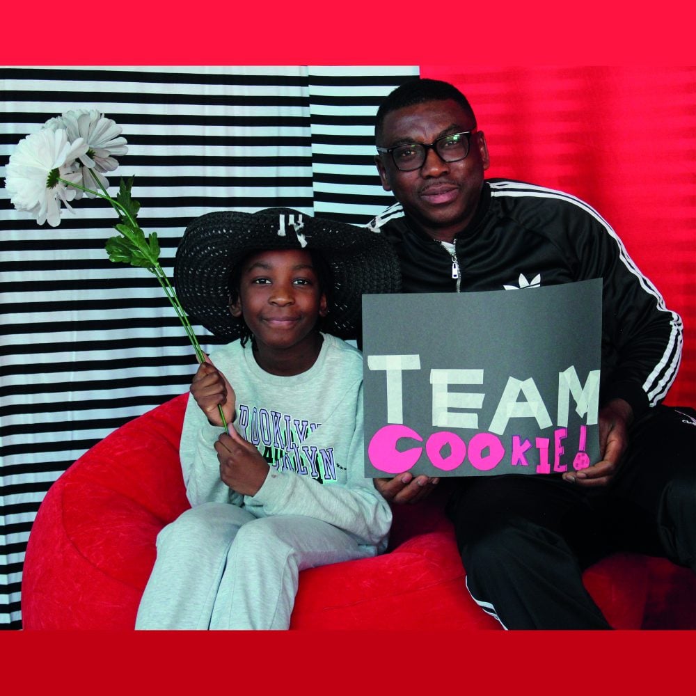 A father holding up a sign with his daughter holding a flower in front of a red and black & white stripe background.