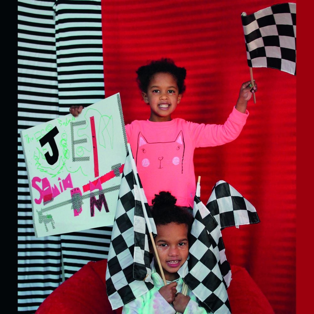 Two children holding up a drawing and three black and white checkered flags on a beanbag in front of a red background.