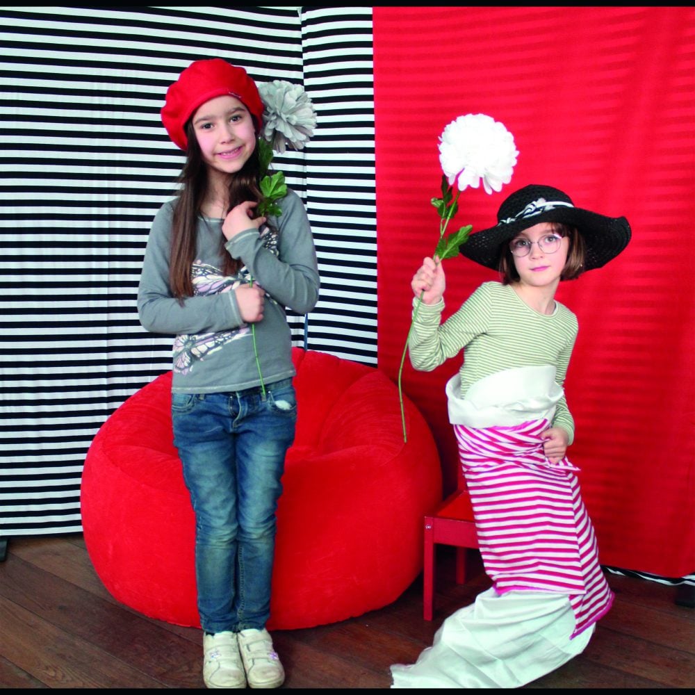 Two girls holding flowers with fancy dress costumes in front of a red and black & white stripe background.