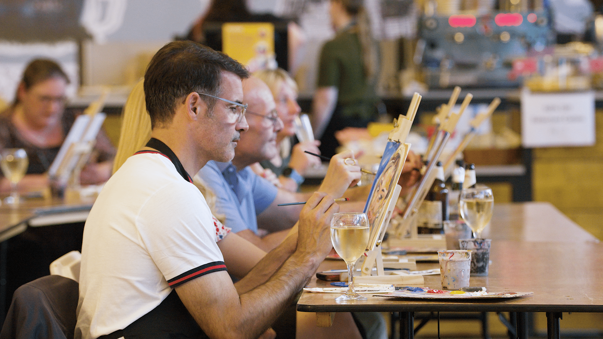people sitting at long tables in the Firstsite foyer painting on small canvases at Boozy Brushes.