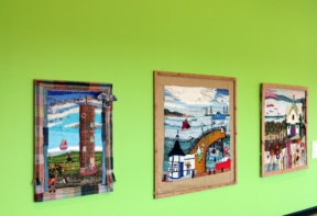 Three framed textile artworks depicting various scenes hang on a bright green wall.