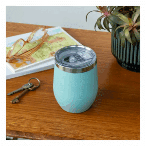 A photo of the blue travel mug sat on a desk along with a plant, some keys, a notepad and glasses