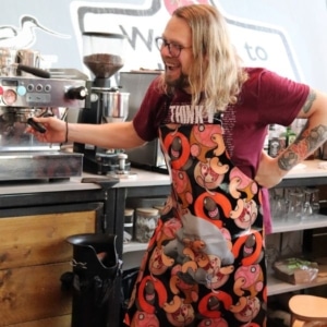 Image of a man stood at a coffee machine wearing a Limied Edition apron designed by Anthea Hamilton.