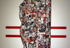 Mixed-media collage shaped like a human torso, suspended by chains against a white wall with three red horizontal lines. The artwork features a dense mix of red, black, and white images, text, and abstract elements.