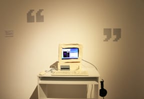 A cardboard off-white colour computer, an off-white painted mouse and keyboard on a table installation. There are headphones and a touchscreen tablet installed inside the cardboard computer.