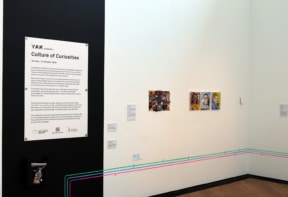 Gallery wall displaying various artworks alongside a sign featuring the exhibition description.