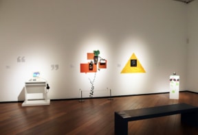 Gallery room with contemporary mixed-media artworks on white walls, a dark bench, and wooden flooring.