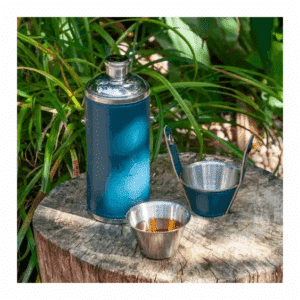 An image of a hip flask sat on a rock with the two included cups of display, one with a golden liquid.