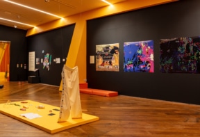Gallery with vibrant orange and black walls displays a variety of contemporary artworks. On the right wall are colourful abstract digital prints. In the centre of the room, a yellow platform holds a sculptural installation featuring oversized white pants standing upright, along with scattered small objects and papers. To the left, mixed-media pieces and a text panel are mounted on the black wall.