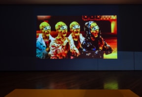 A dark gallery space features a brightly lit video projection on a wall. The projection shows four figures with bright yellow heads and exaggerated cartoonish eyes, wearing different clothing styles.