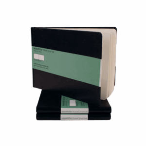 A stack of travel journals showing the front cover. Cover is black, there is a green label giving some information on the product.