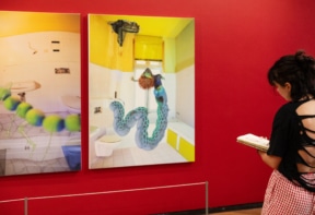 A woman with a sketchbook observes surreal artwork in a gallery with red walls. The art features distorted bathroom scenes with fantastical, snake-like creatures and unusual elements.