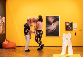 Two people view intimate photographic artworks in a brightly lit gallery with bold yellow walls. Nearby, white trousers with handwritten text are displayed upright, and an orange beanbag sits in the corner.